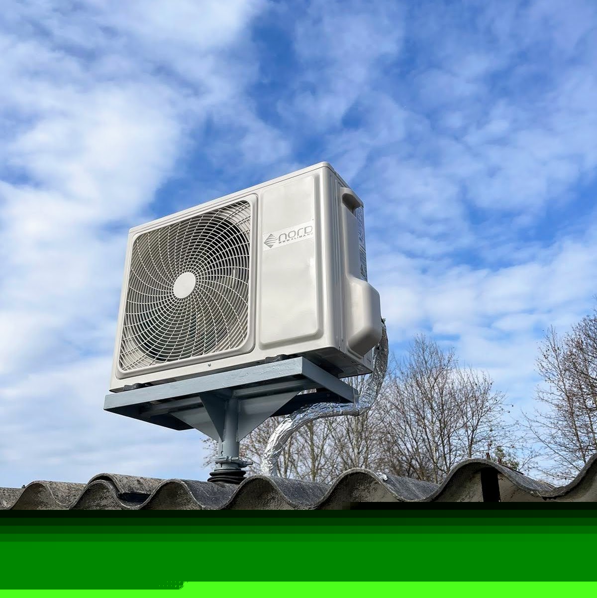 Kültéri klímaberendezés kültéri egysége tetőcserepeken, ventilátorrács látszik kék ég és fák felett. Mohács 